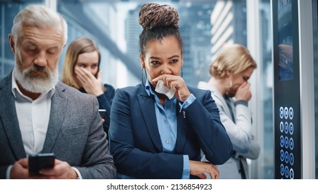 Middle Aged Woman Sneezes In A Crowded Glass Elevator In A Modern Office Building. Businesswoman Covers Her Face, But Other People Are Afraid To Catch The Virus And Microbes In A Lift.