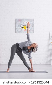 Middle Aged Woman Standing In Triangle Pose While Practicing Yoga On Mat At Home