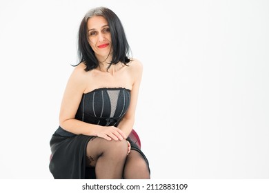 Middle Aged Woman Starting Getting Grey-haired Is Posing In Studio While Sitting On The Chair In Black Dress And Stockings On White Background, Middle Age Skincare Cosmetics, Cosmetology Concept