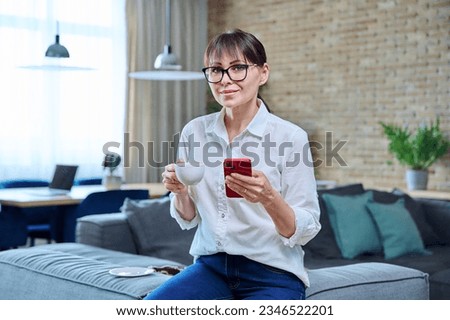 Middle aged woman using smartphone, having cup of coffee at home