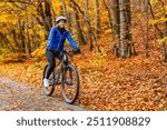 Middle aged woman wearing blue sporty jacket, black cycling pants and white bike helmet riding bicycle in city forest in autumn scenery. Front view.