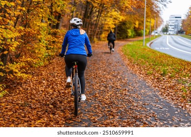 Middle aged woman wearing blue sporty jacket, black cycling pants and white bike helmet riding bicycle on bike path in city in autumn scenery. Back view. - Powered by Shutterstock