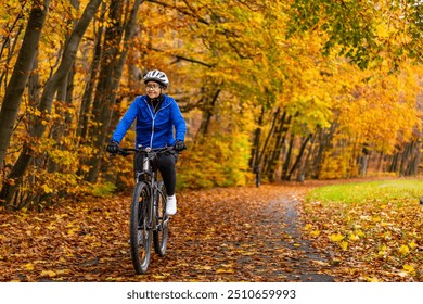 Middle aged woman wearing blue sporty jacket, black cycling pants and white bike helmet riding bicycle in city forest in autumn scenery. Front view. - Powered by Shutterstock