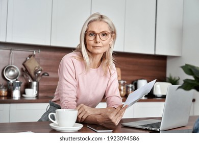 Middle aged woman wearing glasses holding document, letter or bill working on laptop sitting at home office kitchen. Mature lady entrepreneur looking at camera while checking financial information.