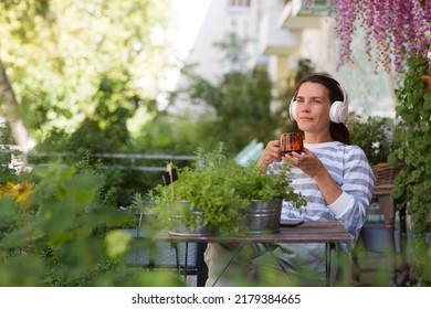 Middle Aged Woman Wearing Headphones With A Cup Of Tea Relaxed In The Garden Near The House In Summer. Concept Without Stress. Mental Health. Slow Life. Enjoying The Little Things. 