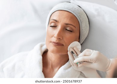 Middle aged woman, wearing medical cap on head, receiving aesthetic facial treatment, beauty specialist giving her anti age injection shot in nasolabial skin fold