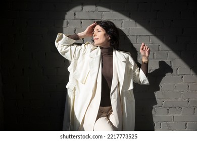 Middle Aged Woman  In A White Cloak Relaxing Against A Gray Wall. Copy Space. 