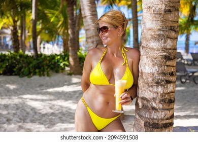 Middle Aged Woman In Yellow Bikini Holding Glass Of Healthy Fresh Fruit Juice On Tropical Beach With Palm Tress