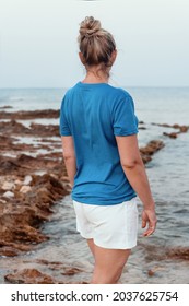 Middle Aged Women In Blue T-shirt With Hair Bun Standing On The Cliff By The Sea At Sunset. Back Shoot, Tshirt Mockup