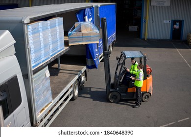 A Middle Aged Worker Driving And Using The Forklift To Load Packages On A Semi-truck Outside The Warehouse With The Help Of A Colleague Standing Beside