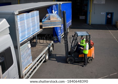 A middle aged worker driving and using the forklift to load packages on a semi-truck outside the warehouse with the help of a colleague standing beside