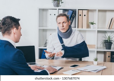 middle aged worker in neck brace with broken arm sitting at table and talking to businessman in blue jacket in office, compensation concept - Shutterstock ID 1307687074