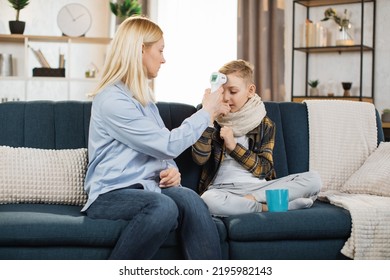 Middle Aged Worried Caring Blond Woman, Mother, Sitting On Sofa At Home, Hugging Her Sick Sad Teen Son, Measuring Temperature Using Infrared Wrist Non-contact Thermometer. Flu, Cold, Virus Infection