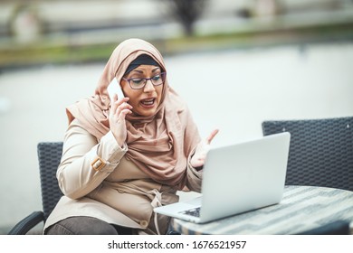 Middle aged worried Muslim woman wearing hijab is sitting on the cafe in urban environment, talking by smartphone and working on laptop. - Shutterstock ID 1676521957