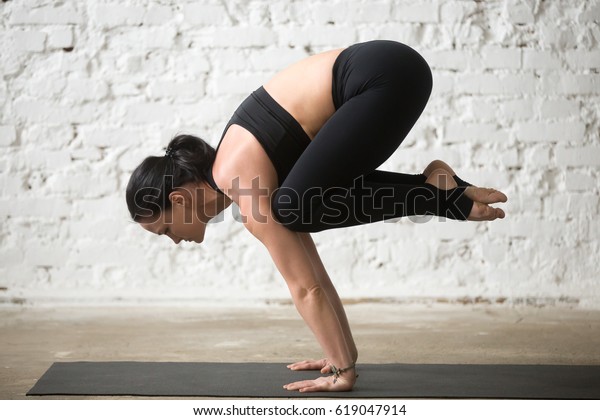 Middle aged yogi attractive woman practicing yoga\
concept, standing in Crane exercise, Bakasana pose, working out,\
wearing sportswear, black tank top and pants, full length, white\
loft background 