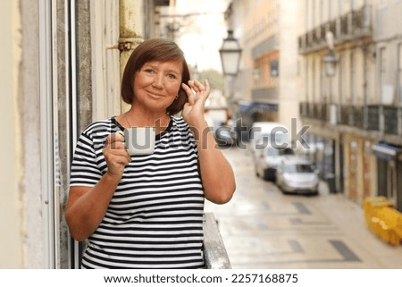 Middle ages woman holds cup of coffee or tea, standing on the balcony with Europe city street view in the morning. Mature female enjoying vacation in resort. Summer, weekend, tourism, travel.