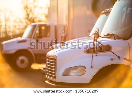 Middle duty rigs semi Trucks with box trailers standing at the gates of the warehouse for loading commercial cargo for the next local deliveries flooded with sunlight