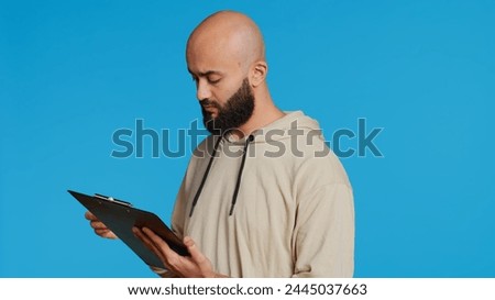 Middle eastern adult creating a checklist on clipboard papers, being focused on trying to plan tasks on an organizer in studio. Modern person looking at files and listing things to do. Camera 2.