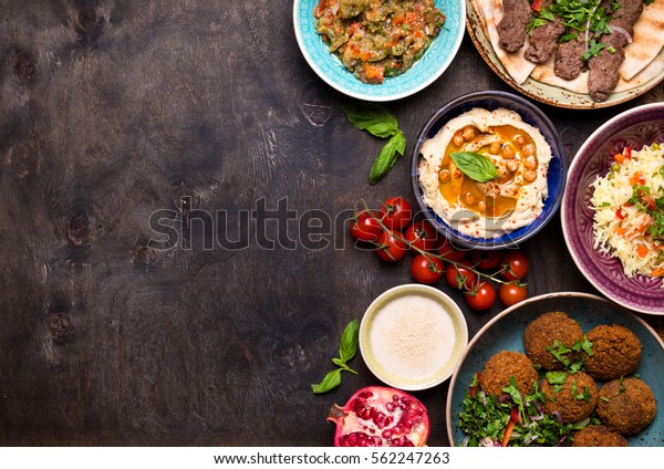 Middle eastern or arabic dishes and assorted meze on\
a dark background. Meat kebab, falafel, baba ghanoush, hummus, rice\
with vegetables, tahini, kibbeh, pita. Halal food. Space for text.\
Top view