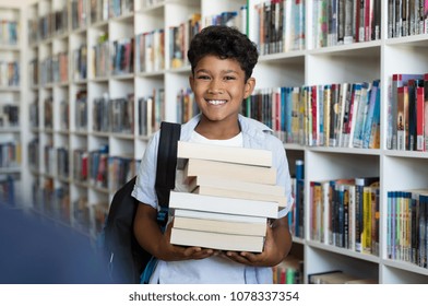 Middle Eastern Boy Holding A Stack Of Books Against Multi Colored Bookshelf In Library. Portrait Of Happy Arab Schoolboy With Backpack At School. Happy Young Indian Child Holding Heap Of Books.