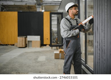 Middle eastern construction worker applying sealant to window frame in prefabricated house factory, ensuring airtight and weatherproof installation