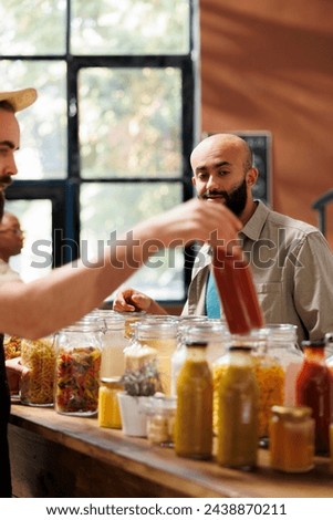 Middle Eastern customer looks at glass bottle filled with organic sauce being held by storekeeper. Local vendor providing male client with bulk natural sustainable products during an event.