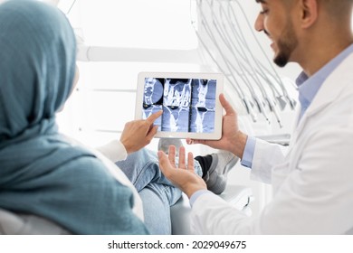 Middle Eastern Dentist Showing Xray Of Teeth Treatment Process On Digital Tablet To Islamic Female Patient During Check Up In Modern Dental Clinic, Muslim Lady In Hijab Pointing At Tab Computer