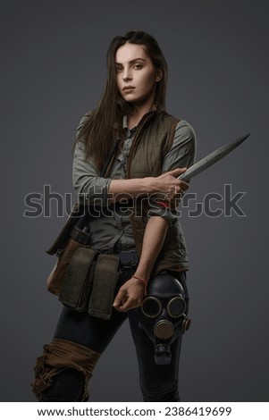 Middle Eastern descent in post-apocalyptic survival gear, brandishing a dagger on a gray backdrop, showcasing her determination to survive
