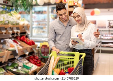 Middle Eastern Family Couple Buying Groceries And Taking Notes In Shopping List Standing And Hugging In Modern Supermarket Indoor. Muslim Spouses Purchasing Food Products In Store