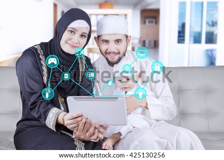 Middle eastern family sitting on the sofa and smiling at the camera while holding a digital tablet with smart house system