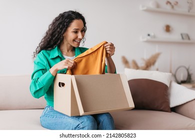 Middle Eastern Female Buyer Holding New Clothes Unpacking Cardboard Box Excited About Successful Shopping Sitting On Sofa At Home. Happy Shopaholic Lady Receiving New Outfit From Shop