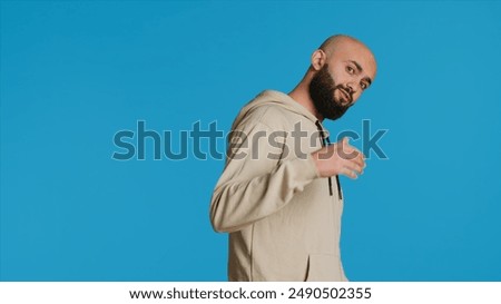 Middle eastern guy asking people to come over, posing over studio background. Arab person invites someone to join him, welcome to come closer on camera, casual invitation. Camera 1.
