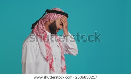 Middle eastern guy dealing with awful headache and tension, suffers from migraine over blue background. Young man wearing national arab costume, feeling sick and stressed with sinus discomfort.