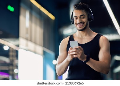 Middle Eastern Male Athlete In Wireless Headphones Using Mobile Phone At Gym, Smiling Muscular Arab Man Relaxing With Smartphone After Sport Training In Fitness Club, Browsing New App, Free Space