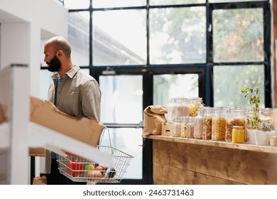 Middle Eastern man browsing different food items in other sections of the store. Male consumer holds basket filled with a mixture of various fruits and vegetables, healthy blend of bio products.
