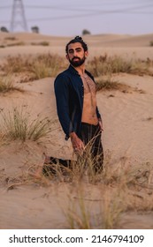 Middle Eastern Man With Curly Long Hair Wearing An Unbuttoned Dark Blue Shirt And Black Skinny Jeans Kneeling In The Desert. Taken In Dubai, United Arab Emirates On August 27, 2021.