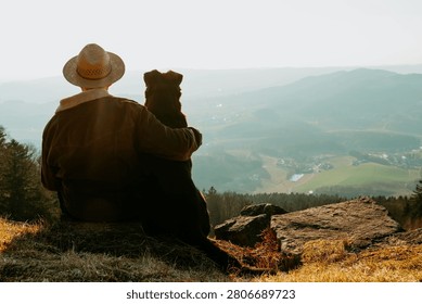 A Middle Eastern Man in a hat sitting with his dog overlooking a scenic mountain valley at sunset in hiking area Bavarian Forest in Germany, Bavaria