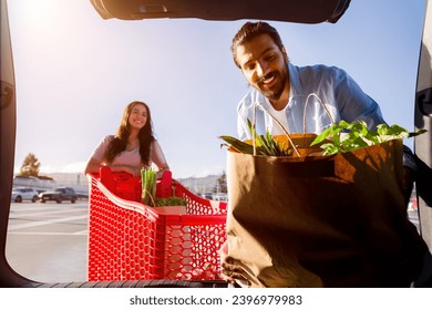 Middle eastern man and woman unloading groceries from a shopping cart into a car trunk, enjoying the sunshine in a supermarket parking lot - Powered by Shutterstock