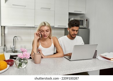 Middle Eastern Man Working And Typing On Laptop While European Girl Talking On Smartphone At Table At Home Kitchen. Young Multiethnic Couple. Remote Work And Freelance. Domestic Lifestyle. Modern Flat