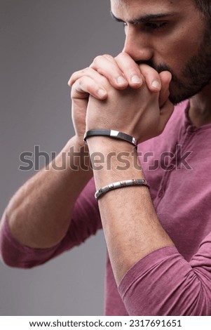 Middle Eastern man's side portrait, hands before mouth and nose as if praying. He's pensive, focused, a well-groomed beard, bracelets, in a maroon shirt. Looking down but not seeming sad.