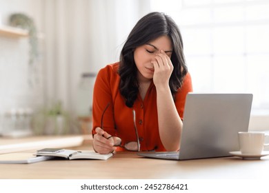 Middle eastern woman appears distressed or exhausted, holding her glasses, with a laptop, notebook, and cup in front of her - Powered by Shutterstock