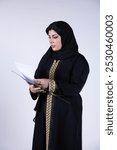 Middle Eastern woman in traditional abaya standing in a white background while holding a holder, checking the paper showing focus on business and professionalism, poised businesswoman