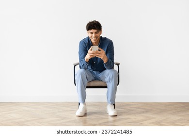 Middle eastern young man texting on his smartphone, smiling enjoying online communication in social media chat, sitting in chair over white wall. Full length shot of guy websurfing and using app - Powered by Shutterstock