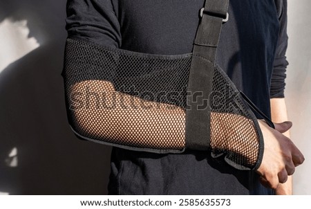 The middle part of the body of a man, whose right arm is wrapped in a black mesh bandage around his neck for support. Medical shoulder elbow support strap, immobilizer brace strap for broken arms.