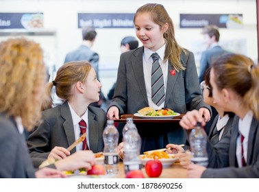 Middle School Girl With A Lunch Tray Standing Next To Her Friends Eating Lunch In The School Cafeteria.