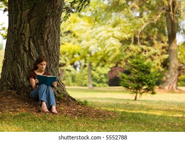 Middle School Girl Reading A Book Under A Large Tree In A Park-like Setting.