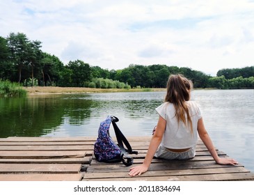 A middle school teenage girl with long hair sits with her back on a wooden pier or pantone and looks at the water. Bank of a river or lake. Next to her is a hiking backpack. Relaxation and tranquility