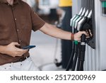 Middle selection of young man holding the phone with one hand and grabbing with the other black fuel pump at self service petrol station for refuelling a car. Diesel 