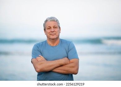 Middle shot of a senior man in blue t-shirt with arms crossed on the chest. Person looking to the camera with a light smile. View against blurry background of sea and sky - Powered by Shutterstock