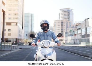 Middle shot of a young motorcyclist stopped at a traffic light in Barcelona. The man rides through the city on his white scooter on a big avenue full of skyscrapers.